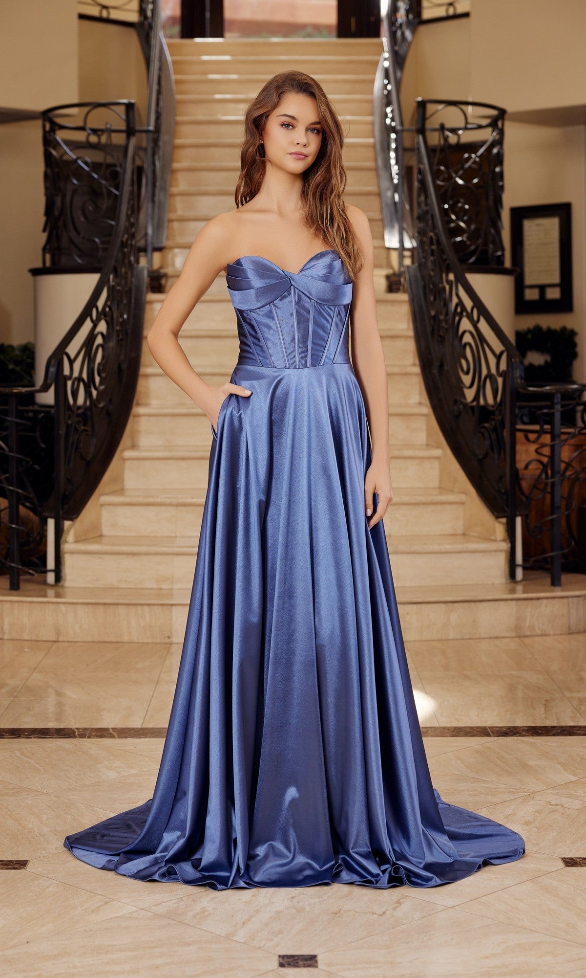 A woman stands on marble flooring before a staircase, wearing the Long Prom Dress: Nox Anabel K1735 in Midnight Blue, which features a strapless satin design with a fitted corset bodice and a flowing, shiny skirt.